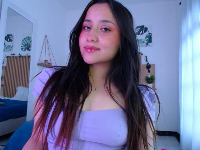 Gabriela Mora mit langen, dunklen Haaren, rosig-roten Wangen, pinkem Lippenstift und einem süßen Lächeln, trägt ein weißes Oberteil, das ihre kleinen Brüste zeigt, in einem hell erleuchteten Raum..