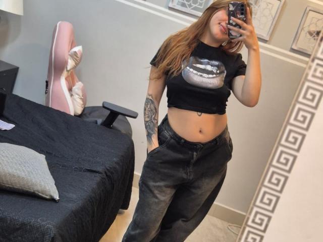MaylinSanchez with long brown hair, black crop top showing her tits, black jeans, tattoo on right arm, taking a mirror selfie, pink gaming chair, and black bed in background.