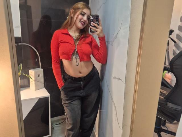 MaylinSanchez with long blonde hair, red cropped top, showing her tits, black pants, hands in pockets, smiling, taking a mirror selfie, holding a phone with a black case.