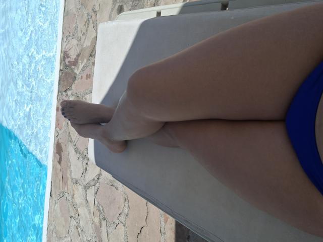 KatherinLatin's legs are crossed on a beige lounge chair by a blue pool, wearing a blue bikini, showing tan skin and her toes with dark painted nails.