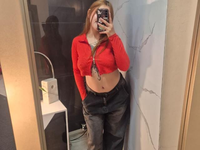 MaylinSanchez with long blonde hair, red cropped sweater, exposing her tits, black pants, standing in front of mirror, taking selfie with phone, reflection on wall, in changing room.