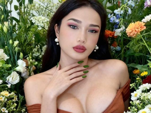 xGoddessJazzyx with green nails, pearl earrings, and auburn dress showing her big tits, surrounded by colorful flowers, looking seductive with pink eyeshadow and red lipstick.