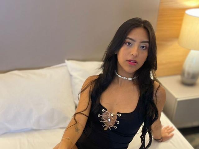 EmmaEstreilla with long black hair, wearing a black dress with metal lace-up detail, silver choker, sitting on a bed, looking at the camera with pink lipstick.