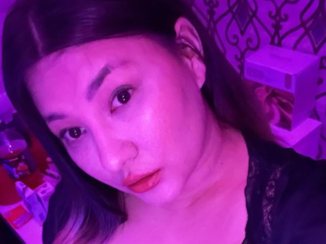 DamelySakura has pink-tinged skin, black lace top, red lipstick, and dark hair in a selfie with purple lighting. Boobs visible, subtle makeup, and a slightly confused expression.