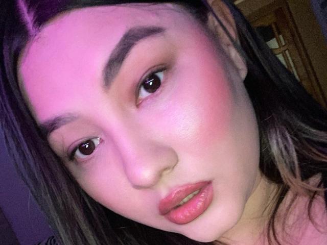 Close-up of DamelySakura's face with pinkish-purple lighting. DamelySakura has light skin, dark brown eyes, and thick, dark eyebrows. Her cheeks are flushed, and her lips are slightly parted with pink lipstick. She has long, dark brown hair with a purple streak. The image highlights her smooth skin texture and subtle makeup. The composition focuses on her face, with her eyes looking slightly to the left. Her expression is neutral, almost pensive. The lighting creates a soft, warm glow on her skin.