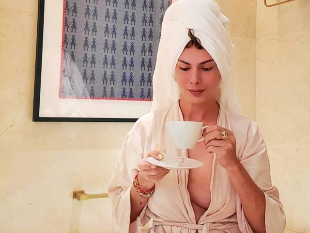 BeautyQueenTs holds a white teacup on a saucer, sipping with a relaxed expression. She wears a beige robe and a white towel wrapped around her head. Her brown hair peeks out. She has gold jewelry on her right hand and a bracelet on her left. A framed patterned artwork hangs in the background. The image is warm-toned with a soft, almost creamy texture. She looks calm and composed.