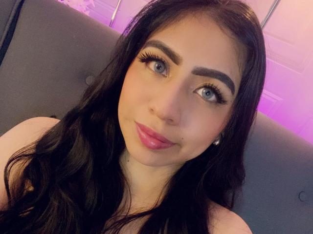 ZoeRedds a de longs cheveux noirs, des yeux bleus, et un rouge à lèvres rose avec de longs cils, elle sourit doucement à la caméra, avec un maquillage subtil qui met en valeur ses traits..