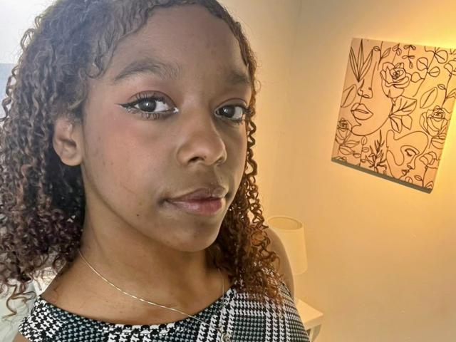 SerenaHottieLady with curly brown hair, black-and-white checkered top, eyeliner, and subtle smile, wearing a thin necklace, standing in a softly lit room. Artwork on the wall.