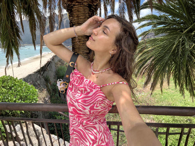 HolaBola with tan skin and brown hair, wearing a pink zebra-striped dress, selfie with eyes closed, touching face, palm tree and ocean in background, bracelet and necklace.