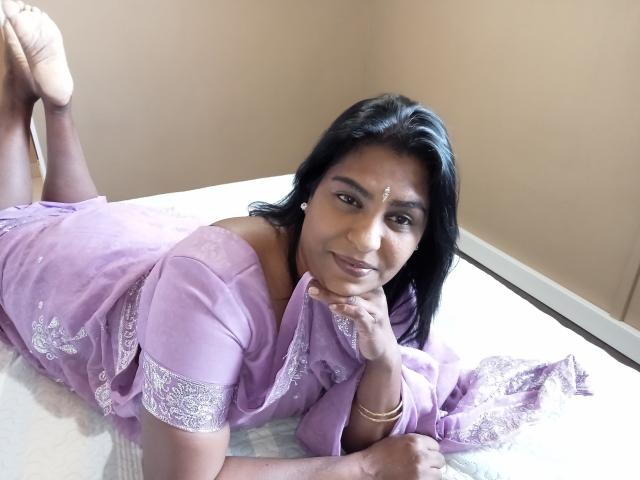 Aadhya è sdraiata su un letto, indossa un sari viola con pizzo bianco, i suoi capelli scuri sono sciolti, sorride, con i glutei sollevati e i piedi in aria..