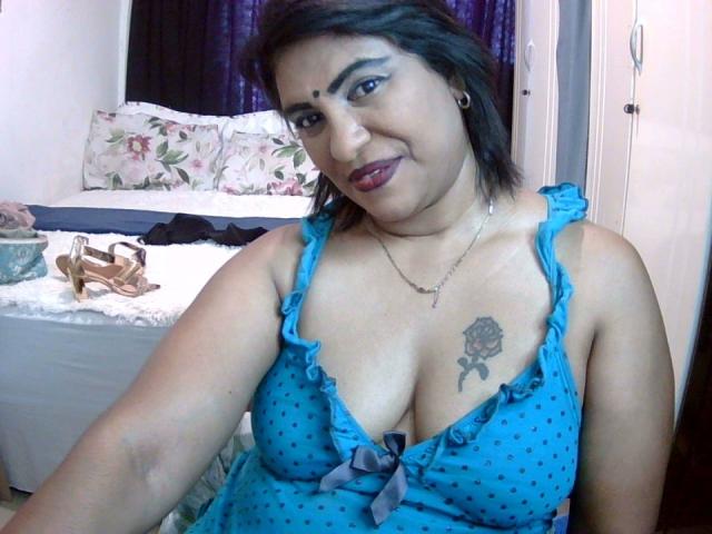 Aadhya, con cabello oscuro, vestido azul con lunares, mostrando sus pechos, tatuaje de rosa en el pecho, lápiz labial oscuro, sonriendo, en el dormitorio, con aros de oro, cortinas moradas en el fondo..