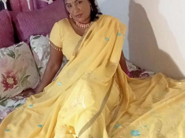 Aadhya, vestida con un sari amarillo con motivos florales azules, está sentada en una cama. Tiene senos de tamaño medio, cabello oscuro y un collar rosa, y mira ligeramente hacia un lado..