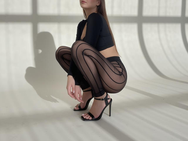 ReginaWein squats in black sheer pantyhose, high heels, and a black crop top, showing her ass and tits. Shadow of her head is visible on the white floor.