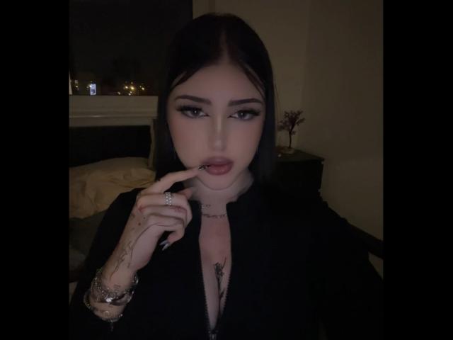 MiaHaveFun with pale skin, dark hair, and heavy makeup, biting her finger, showing tattoos, in a black shirt, with a ring and bracelet, in a dimly lit room.