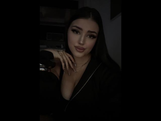 MiaHaveFun with dark hair, full lips, and serious expression, wearing a black robe, showing her cleavage, hand on chin, and a gold necklace, in dimly lit room.