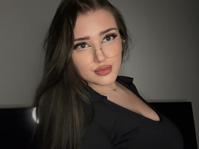 MiaHaveFun has long, straight brown hair and is wearing thin, gold-rimmed glasses. She has light skin and full lips with a slight pout. She's wearing a black V-neck shirt that highlights her chest. Her eyebrows are dark and well-defined. The lighting is even, showing her clear, smooth skin. The image focuses on her upper body and face. Her expression is neutral, with a slight, almost imperceptible smile. The overall composition is simple and centered on her face.