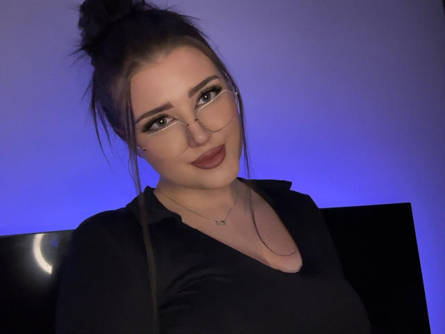 MiaHaveFun with glasses, dark hair in a bun, black shirt, and lipstick, smiling at camera, blue light in background. Her tits are slightly visible under the shirt.