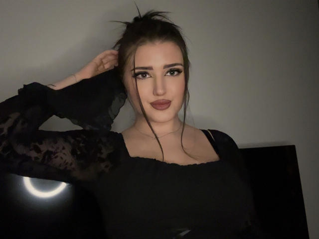 MiaHaveFun with dark hair in a messy updo, wearing a black lace top, has large boobs, and is looking at the camera with a slight smile.