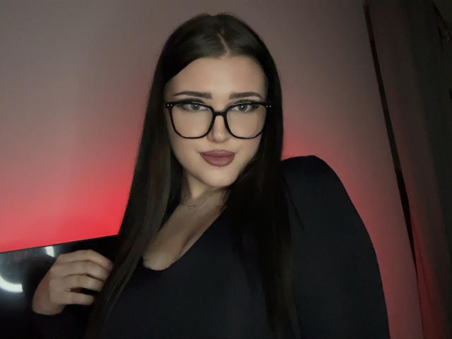 MiaHaveFun with long brown hair, black glasses, and dark lipstick, wearing a black top, has her hand on her chest, looking at the camera with a slight smile.