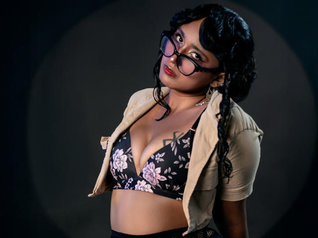 AmelieRose with curly black hair, glasses, red lipstick, beige shirt, black floral bra, showing her tits, dark background, looking up, slightly leaning to the side.