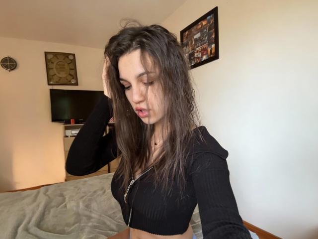 HolaBola with long brown hair, black ribbed crop top, hand in hair, looking down, showing her small tits, in a bedroom with a flat-screen TV and wall art.