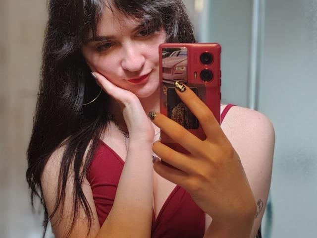 BerryDiva with long dark hair, red dress, holding red phone, smiling, hand on cheek, gold nail polish, hoop earrings, visible tattoo on left forearm, mirror selfie.