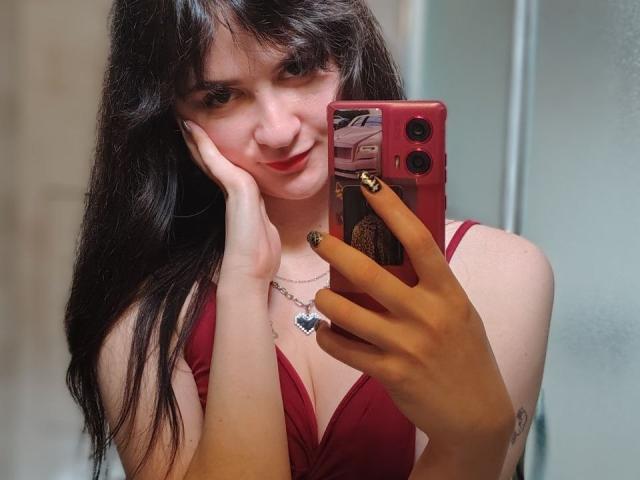 BerryDiva with long black hair, red dress, holding a red phone, smiling, hand on cheek, heart necklace, gold nail polish, mirror selfie, showing her tits.