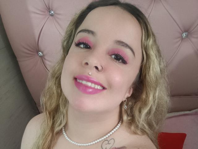 GwenDolenAlana with pink eyeshadow, glossy pink lipstick, nose ring, pearl necklace, and wavy blonde hair, smiling at the camera. She's got a heart tattoo on her chest.