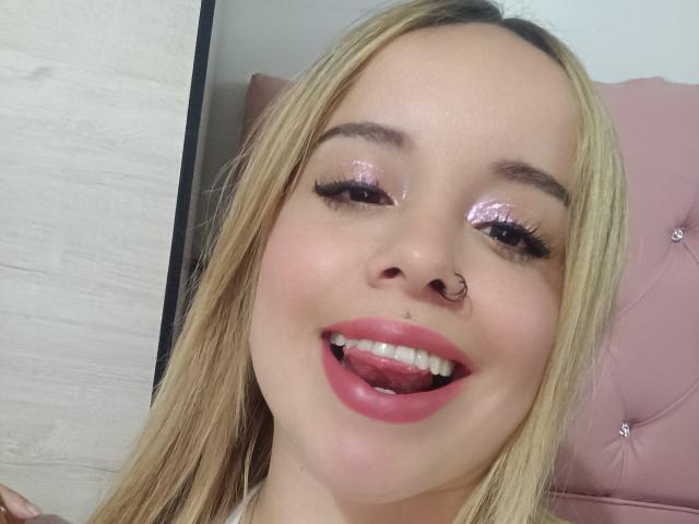 GwenDolenAlana with blonde hair, pink glitter eyeshadow, nose ring, pink lipstick, tongue out, showing teeth, and long eyelashes, against a pink tufted headboard.