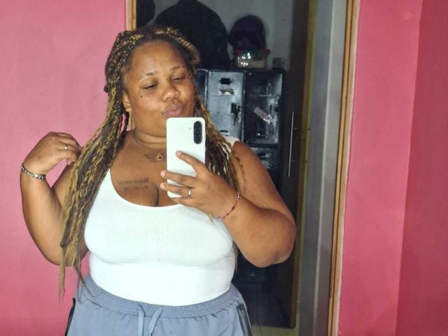 A plus-sized, black woman with long, braided hair takes a mirror selfie, pouting her lips, wearing a white tank top and gray pants, holding a white phone.