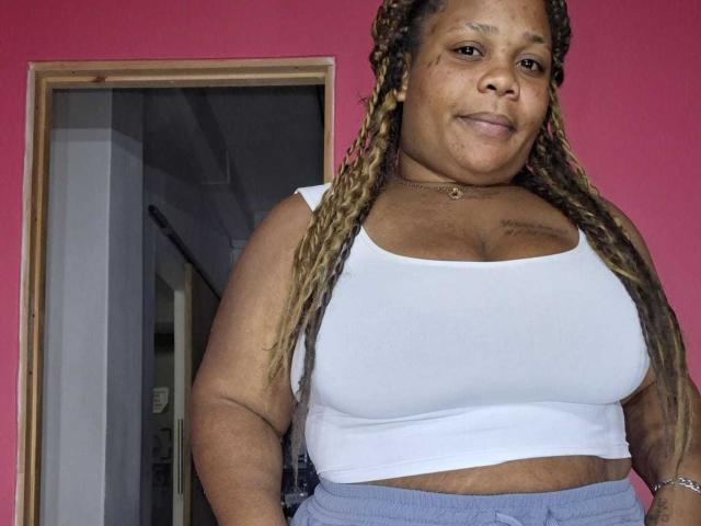 A plus-sized woman with brown skin and long braided hair stands in a pink room, wearing a tight white crop top that shows her big tits and gray pants.