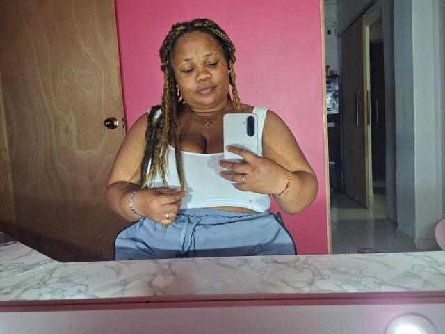 Likechocolate, a plus-sized woman with brown skin and braided hair, takes a selfie in a mirror wearing a white tank top and blue pants, showing her big boobs.