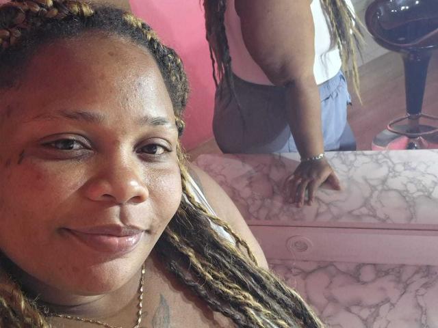 A photo of likechocolate with dark brown skin and braided hair, showing a slight smile, in front of a pink marble countertop with another likechocolate in the background.