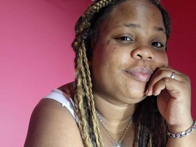 A photo of likechocolate with braided hair, brown skin, and a small smile, resting her chin on her hand, showing a ring and a bracelet against a pink background.