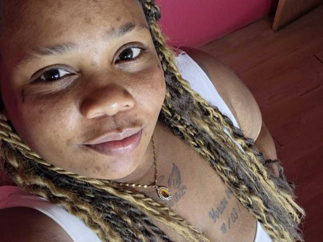 Likechocolate has brown skin, braided blonde and brown hair, wearing a white tank top, smiling, and has tattoos on her chest. She's wearing a gold necklace with a yellow gem.