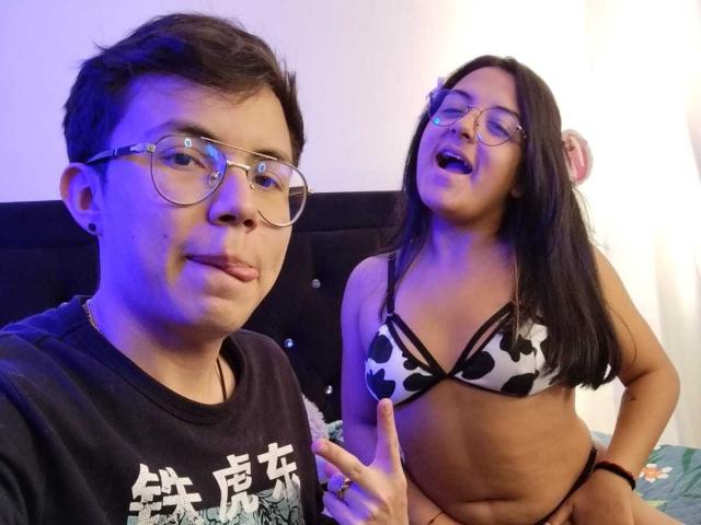 TinnaCharlie with glasses, black shirt, short hair, and slight smile, stands next to TinnaCharlie in a cow print bikini, laughing, with blue light on left. Both have medium skin.