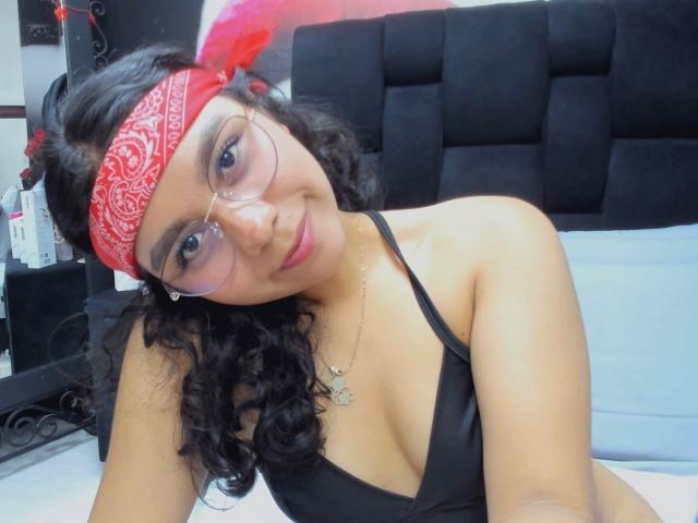 JuliaBennett with curly black hair, red bandana, glasses, black tank top, and necklace, showing small tits and smiling, in a bedroom.