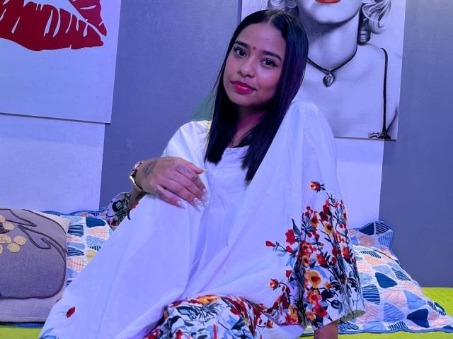 KiaraPriya with straight black hair, wearing a white dress with colorful floral patterns, sitting on a bed, smiling, red lipstick, bracelet on left wrist, looking relaxed.