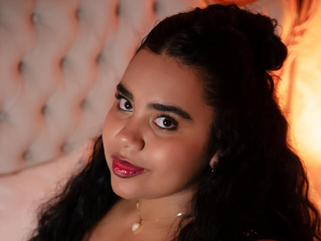 LisaTobon has dark curly hair in a bun, brown eyes, pink lipstick, and a soft smile. She's wearing a gold necklace with a small pendant. Her ass is not visible.