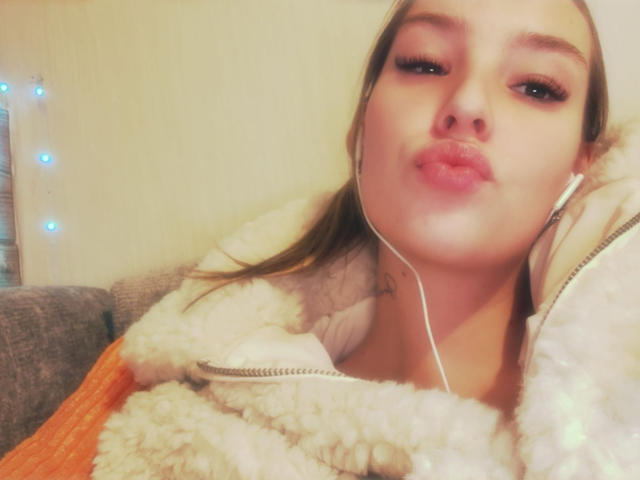 AngelaLuxe with pouty lips, wearing a fluffy jacket, white earbuds in, brown hair, and a tattoo on her neck, looking into the camera with half-closed eyes.