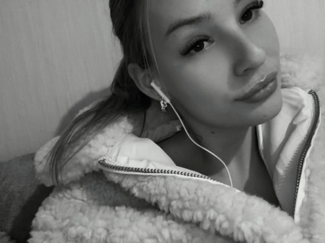 AngelaLuxe with pouty lips, wearing a fuzzy jacket, earbuds in, and her ponytail over her shoulder, in a black-and-white selfie. Her face is slightly tilted to the right.