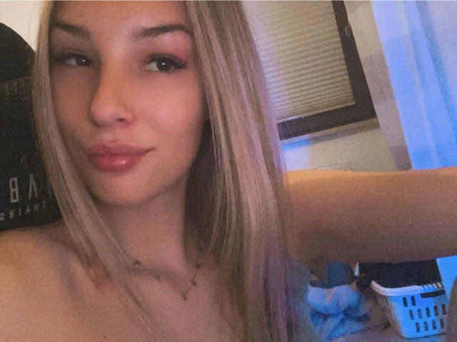 AngelaLuxe with blonde hair, pink lipstick, and a slight pout, showing her bare shoulders, with a necklace, in a soft-lit room, arm raised. Her tits are not visible.