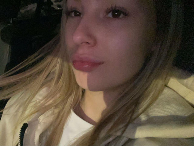 AngelaLuxe's face, with shiny pink lips, blonde hair, and a light jacket, in a dimly lit photo. Her eyes are slightly open, and she's giving a subtle smile.