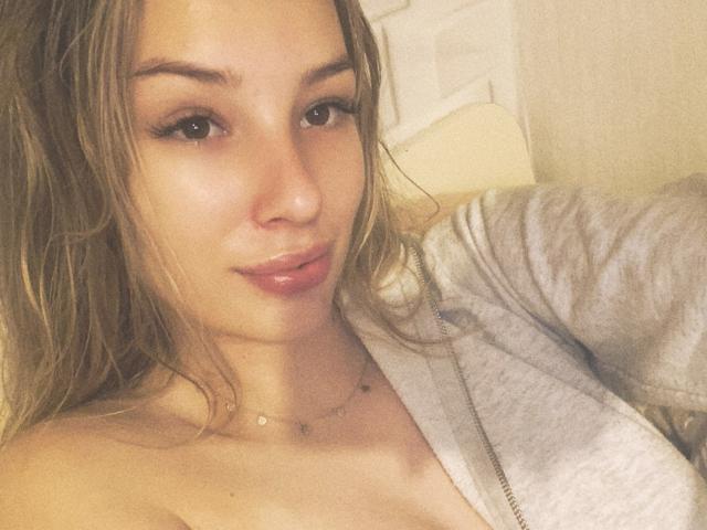 AngelaLuxe with wet, wavy blonde hair, full pink lips, and a gray zip-up hoodie, showing her bare shoulders and small tits. She's wearing a delicate necklace.