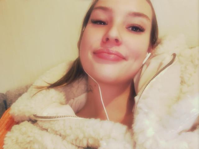 AngelaLuxe with a cute smile, wearing a fluffy white jacket, earbuds in, brown hair, and glossy pink lips, looking slightly up. Soft lighting.