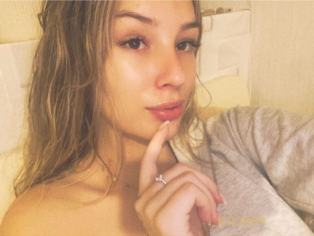 AngelaLuxe has wet, wavy brown hair, full lips, and is touching her chin with her finger, wearing a gray top, showing her ring and subtle makeup.