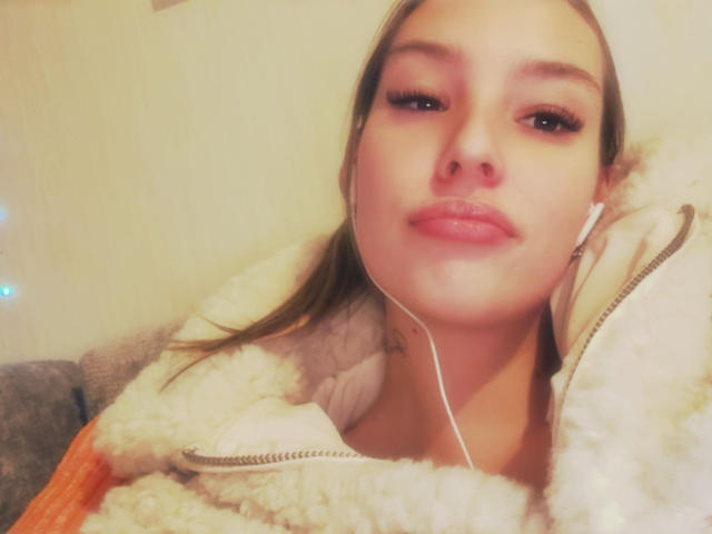 AngelaLuxe with pink lipstick, brown eyes, and white earbuds, wearing a fluffy beige jacket, has a slight smile and her brown hair is pulled back.