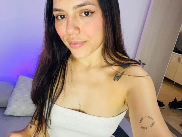 SophiaMarx with long dark hair, white tube top, small tits, tattoos on arm and shoulder, pinkish skin, smiling, brown eyes, in a well-lit room, purple light in background.