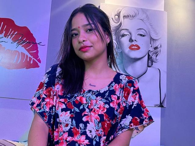 KiaraPriya with medium tits, dark hair, pink lipstick, wearing a floral blue dress, standing in front of black-and-white Marilyn Monroe and red lips paintings.