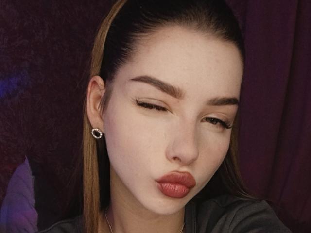 BitterPeach has fair skin, brown hair in a ponytail, and is puckering her red lips while winking. She's wearing a green top and small round earrings.